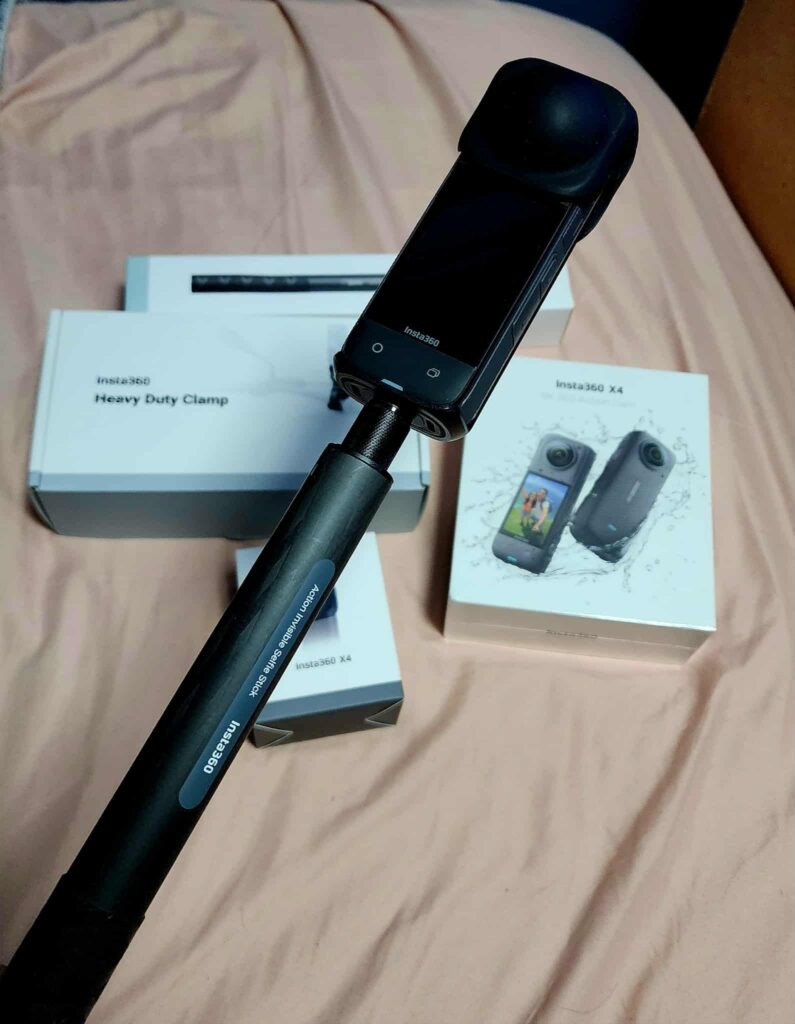 Insta 360 X4 camera mounted on a selfie stick, shown with original packaging including heavy duty clamp and lens cap.
